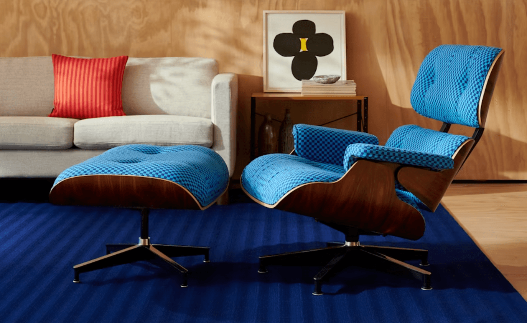 Ghế Eames Lounge Chair và Ottoman màu xanh dương nổi bật với khung gỗ tối màu trên thảm xanh sọc, đặt trong không gian phòng khách hiện đại.