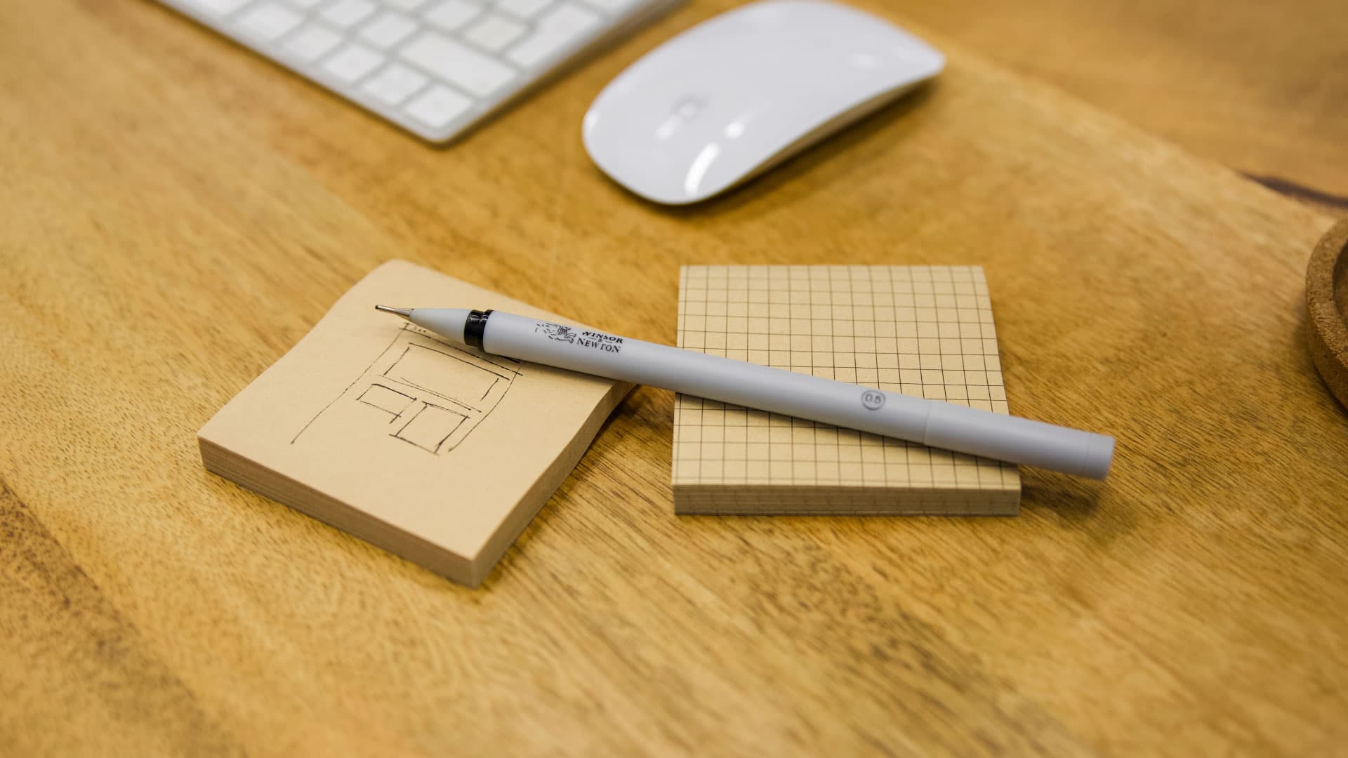 Bút fineliner màu xám đặt trên hai chồng giấy ghi chú màu nâu với bản phác thảo kiến trúc.