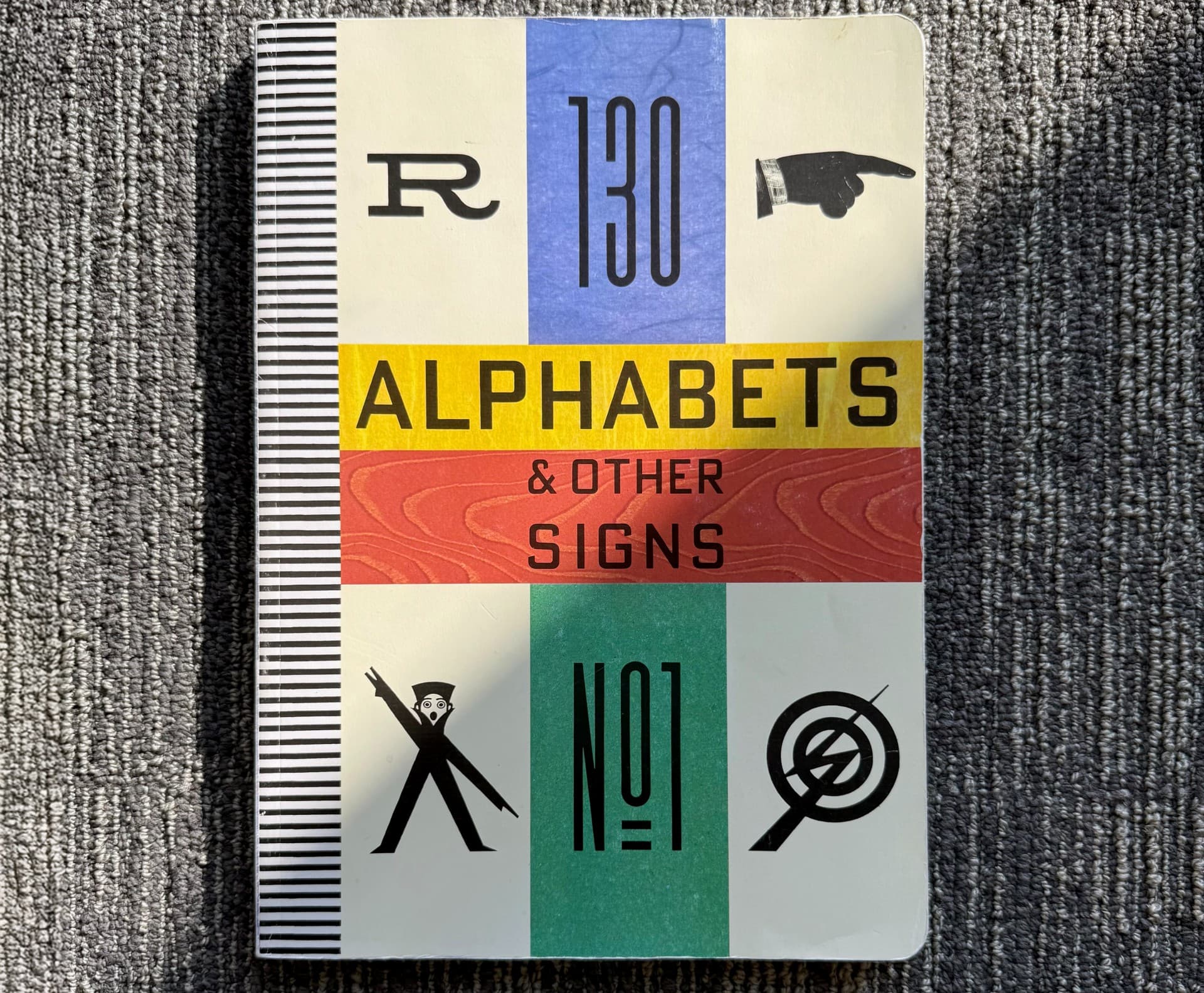 Bìa cuốn sách "130 Alphabets & Other Signs" với các sọc màu rực rỡ và ký hiệu đồ họa.