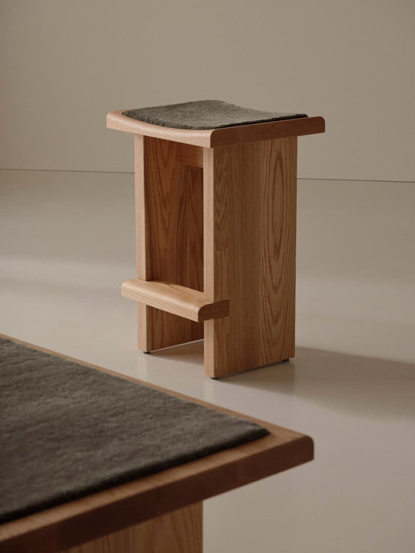 A minimalist wooden stool with a flat gray cushion sits on a smooth, light-colored floor. Part of a matching bench is visible in the foreground.