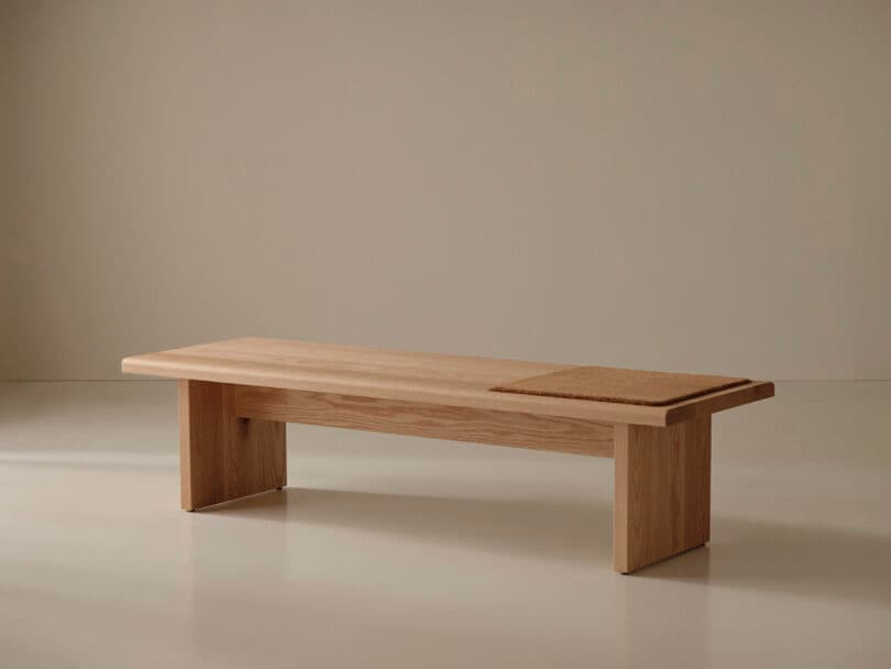 A minimalist wooden bench with a rectangular seat and sturdy legs, featuring a section with a woven or textured surface on one end, set against a plain background.
