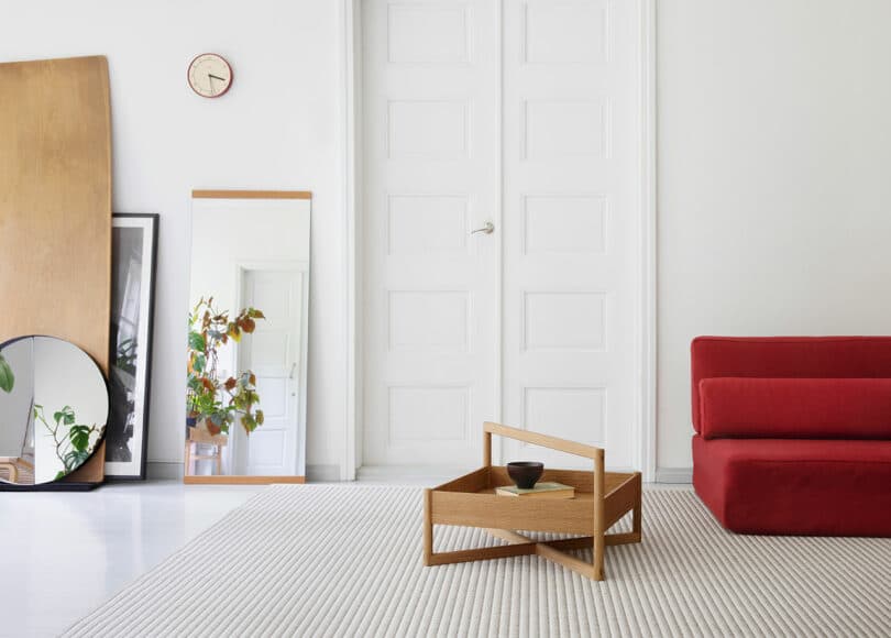 Phòng khách tối giản với ghế sofa đỏ, bàn cà phê gỗ, gương và đồng hồ trên tường trắng, cửa lớn màu trắng và chậu cây